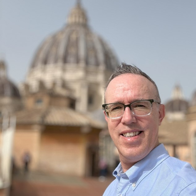 Man wearing glasses taking a selfie with a large domed building in the background.
